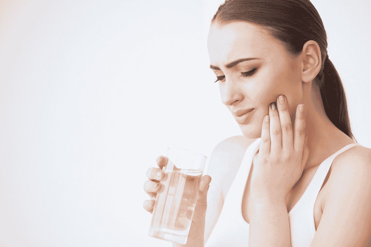 why tooth hurts drinking cold water Woman holding a glass of water and touching her cheek in discomfort from sudden cold tooth sensitivity.